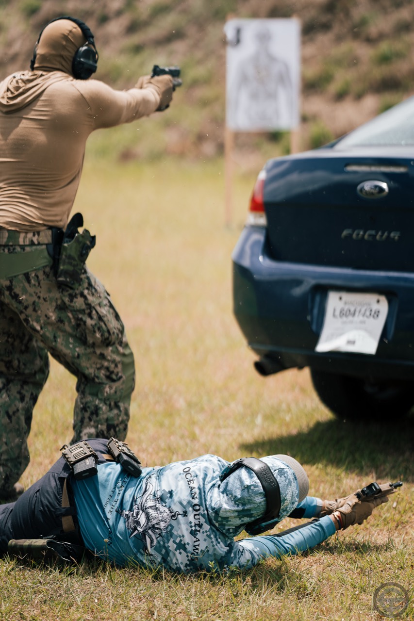 Two shooters engaging paper targets from standing and prone positions near a vehicle